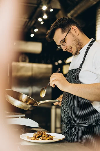 Un chef cuisinier souriant en tablier dressant avec soin un plat gastronomique chaud dans une cuisine professionnelle.